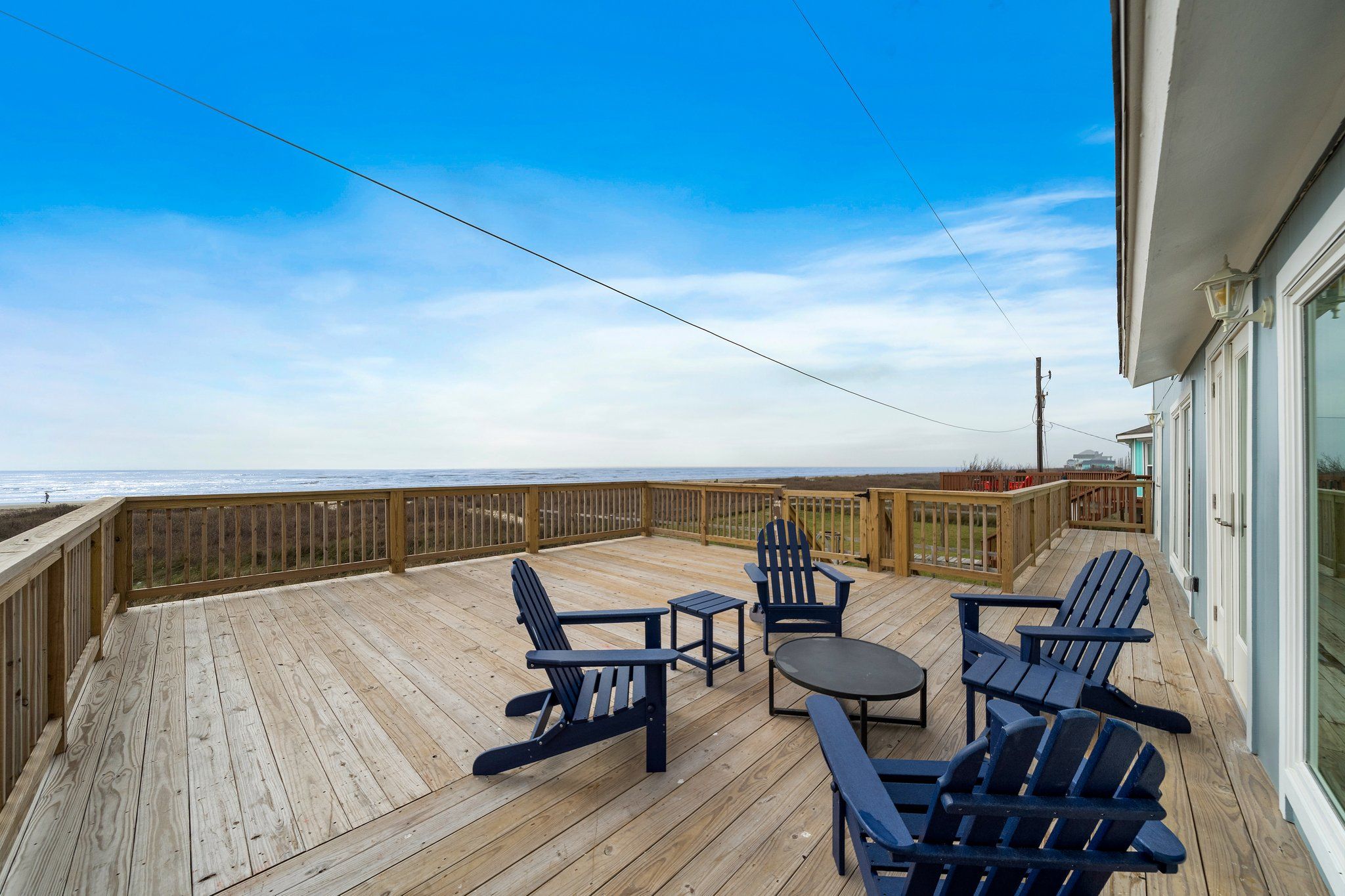 Una terraza de madera con sillas Adirondack y una pequeña mesa, con vista a la playa.