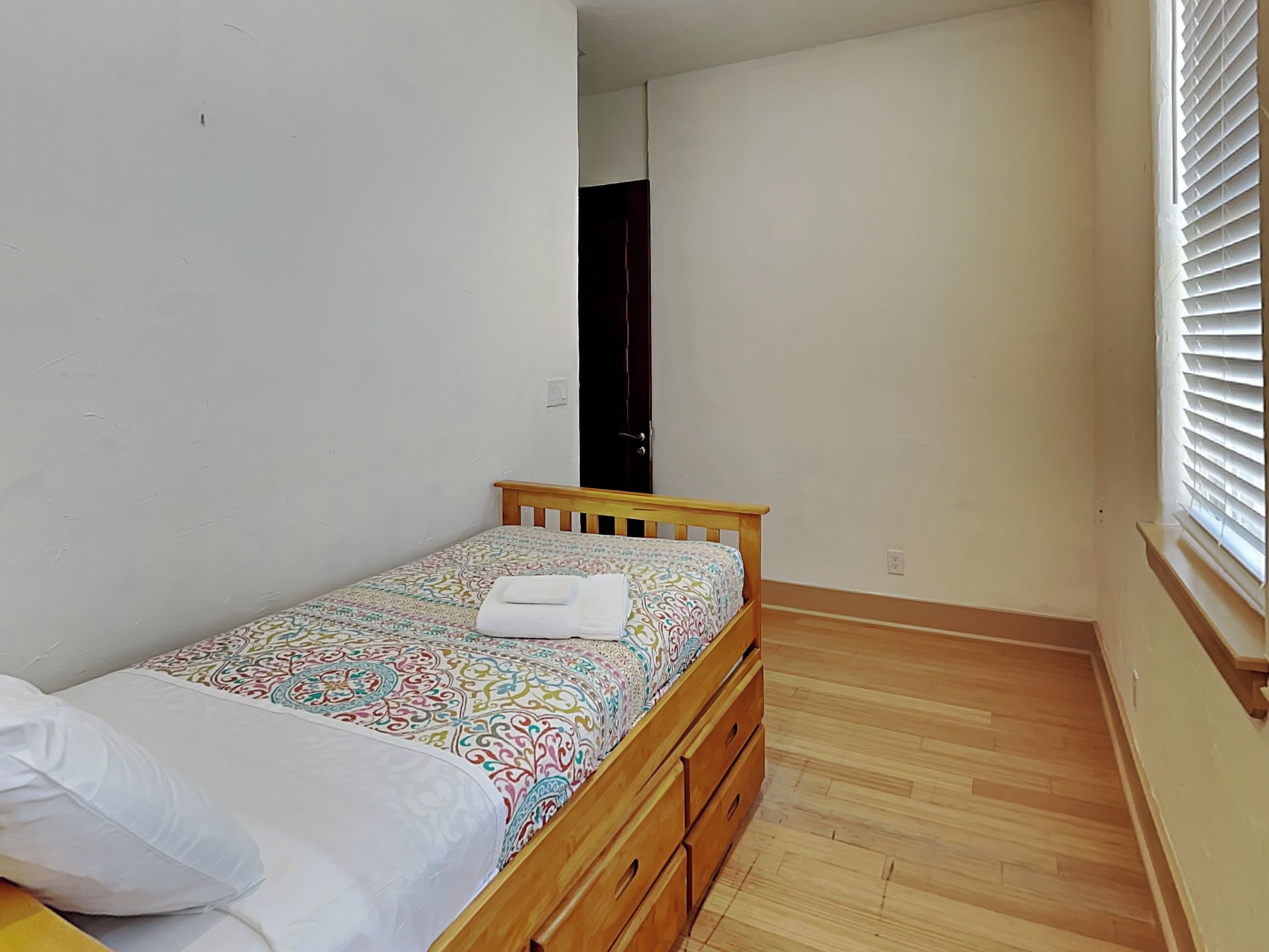 A small, single-bed room with a wooden bed frame, a patterned bedspread, and a white pillow. There is a wooden dresser with drawers, a door in the background, and a window with blinds.