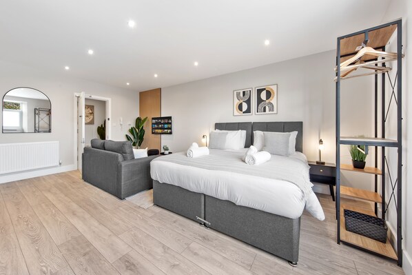 Un dormitorio moderno con una cama grande, un sofá gris, un televisor y piso de madera.