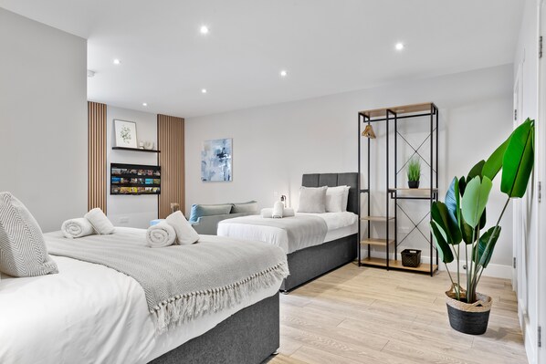 Une chambre moderne avec un grand lit, un plus petit lit, un canapé gris, une télévision, une bibliothèque et une plante en pot.