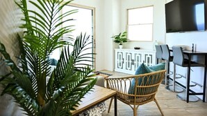Una sala de estar moderna con una silla de mimbre, una mesa de centro de madera y una planta grande en maceta. Hay un comedor con un armario blanco y un televisor montado en la pared.