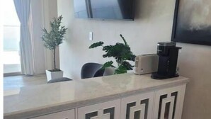 Una sala de estar moderna con un mueble blanco, un televisor de pantalla plana montado en la pared, una cafetera y dos plantas en macetas.
