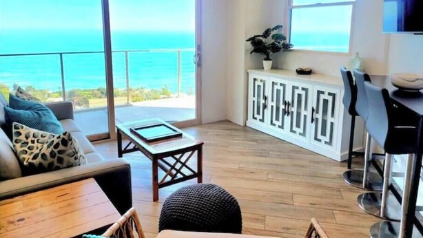 Una sala de estar moderna con vista al océano, un sofá, una mesa de centro y una zona de comedor con taburetes de bar.