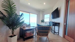 Un salón amplio con una planta grande en maceta, una silla de mimbre, una mesa de comedor y un televisor montado en la pared.