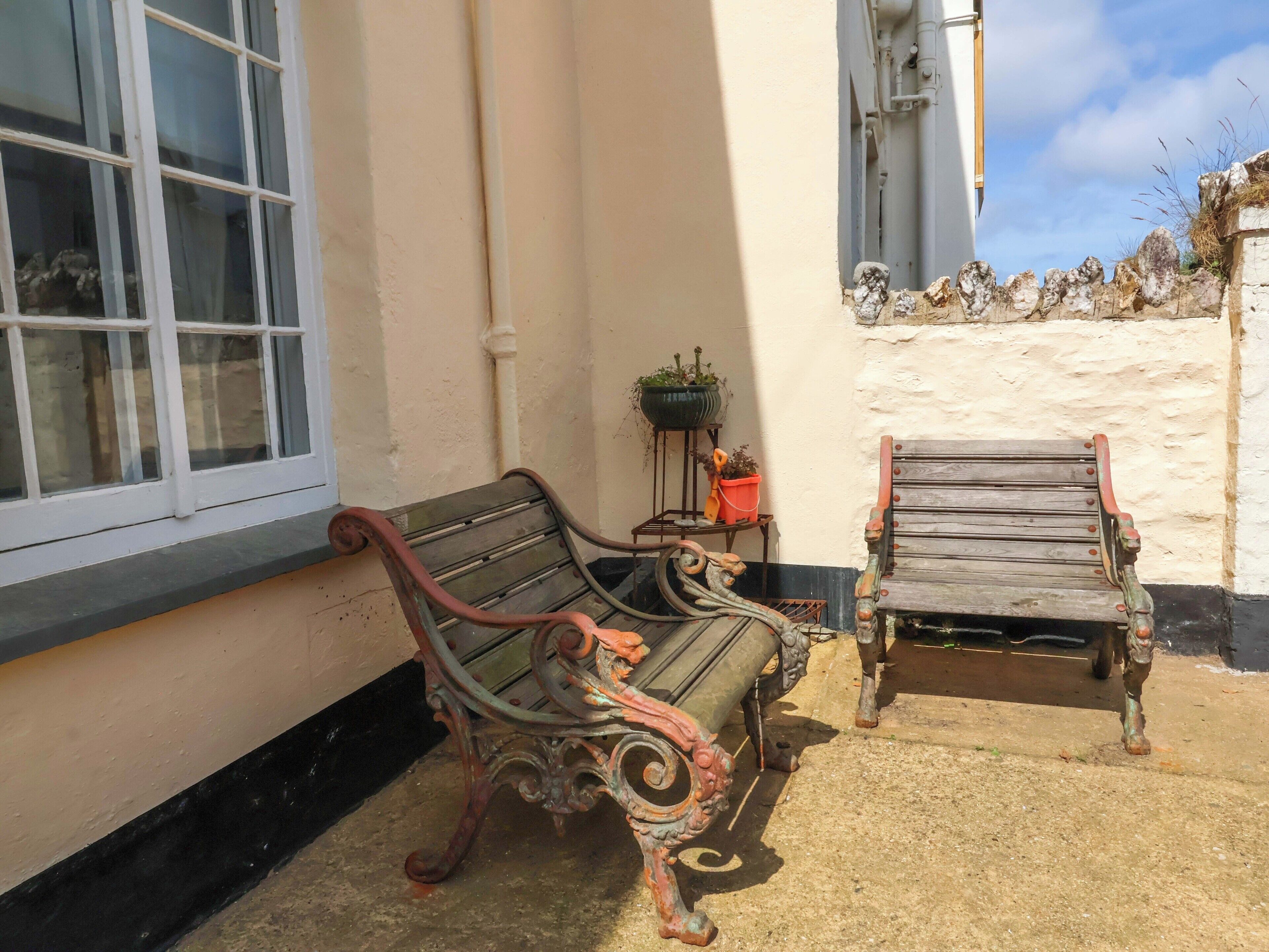 A bench and a wooden chair outside a building with a window and a wall-mounted planter.