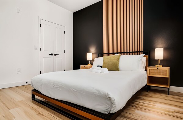 Une chambre d’hôtel moderne avec un grand lit, un plancher en bois, des murs noirs et blancs, et deux tables de chevet avec des lampes.