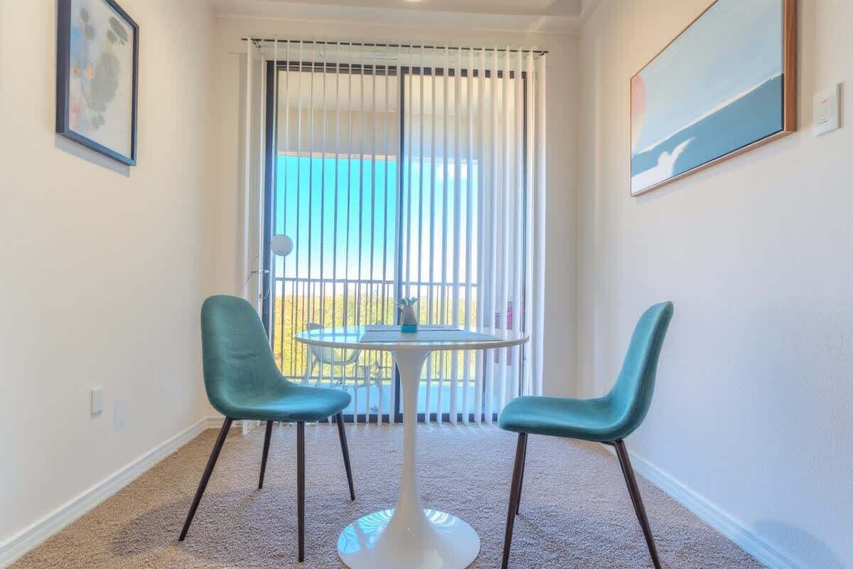 Une pièce avec une table ronde et deux chaises, une fenêtre avec des stores et un tableau encadr é au mur.