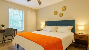 A hotel room with a bed, two bedside tables, a desk, a chair, a ceiling fan, and a window with blinds. - Boulevard Stays Calm 1BR King Retreat Poolside Bliss Gym & Parking (Arlington)
