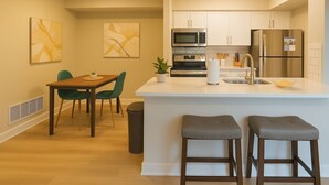 A modern kitchen with white cabinets, a stainless steel microwave, and a refrigerator. There is a dining area with a wooden table and green chairs. - Boulevard Stays Calm 1BR King Retreat Poolside Bliss Gym & Parking (Arlington)