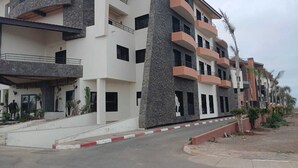 A multi-story residential building with balconies, a central stone pillar, and a paved entrance area.