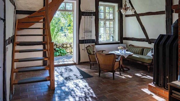 Una habitación acogedora con una escalera de madera, una pared de piedra, una ventana con cortinas, una mesa con sillas y un sofá verde.
