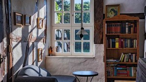Una habitación acogedora con un ventanal grande, una estantería, una mesita redonda y un sillón con cojines.
