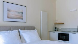 Una cama bien tendida con sábanas blancas en una habitación moderna con cabecera gris, una lámpara colgante, una zona de cocina con microondas y un cuadro en la pared.