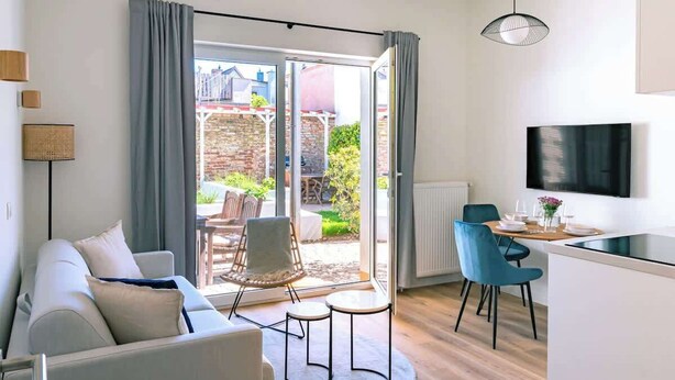 A modern living room with a sofa, a dining area with chairs, a kitchen with a TV, and a sliding glass door leading to a patio. - 2-Raum Appartement „Nr. 3“ mit großer Terrasse (Rostock)