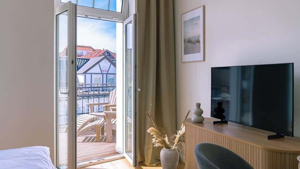 Habitación de hotel con un gran ventanal corredero que da a un balcón, un televisor de pantalla plana sobre un mueble de madera y una cama con colcha blanca.
