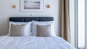 Una cama bien hecha con ropa de cama blanca y beige, un cuadro en la pared y una ventana con cortinas.