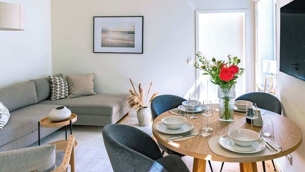 Un coin repas avec une table ronde en bois, dressée pour quatre personnes, entourée de chaises à revêtement gris. Un canapé gris et une petite table d’appoint avec un vase de fleurs rouges sont visibles à l’arrière-plan.