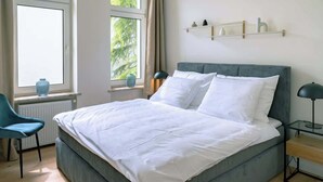 Ein Schlafzimmer mit einem Bett, einem Sessel, einem Nachttisch, einem Fenster und einem Regal mit Dekoration.
