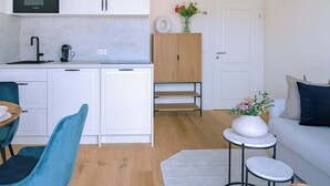 Eine moderne Küche mit weißen Schränken, einer schwarzen Mikrowelle und einer Kaffeemaschine. Es gibt einen Essbereich mit einem blauen Stuhl und einem kleinen Tisch. Im Wohnbereich befindet sich eine weiße Couch und ein runder Tisch mit einem Blumenstrauß.