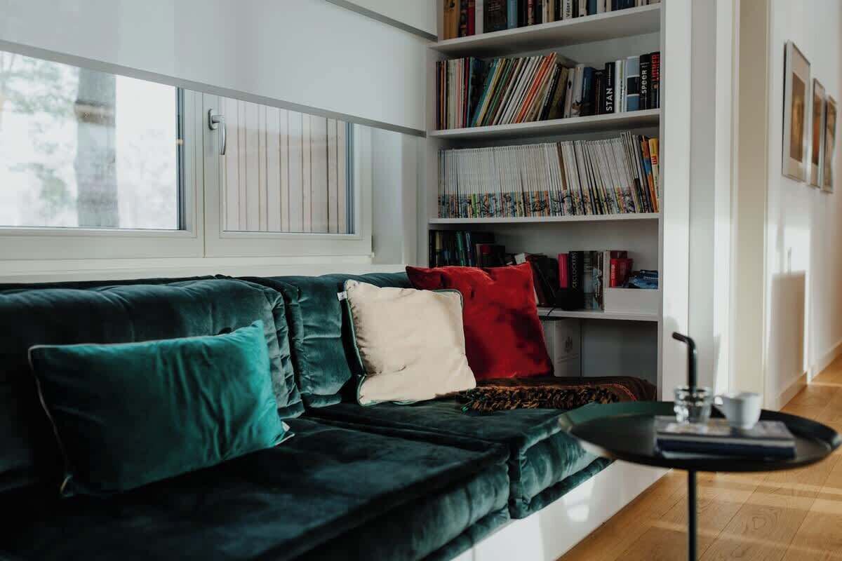 A modern living room with a dark green velvet sofa, a bookshelf filled with books, and a window with blinds.
