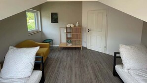 A small, sloped-roof room with a wooden floor, a yellow chair, a white chair, a wooden shelf unit, and a white door. - "Smart Apartment" Heidelberg (Heidelberg)