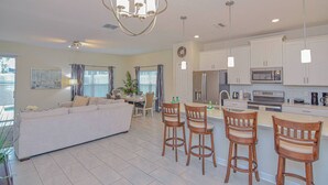 A modern kitchen with white cabinets, a central island with bar stools, and a dining area with a white sofa and table. - Go 27 - Champions Gate (Kissimmee)