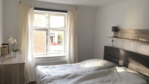 Une chambre à coucher avec un lit, une fenêtre avec des rideaux, une table de chevet avec une plante et une étagère fixée au mur.
