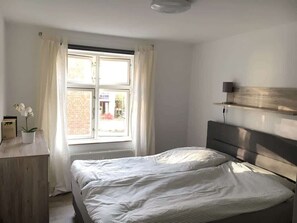 A bedroom with a bed, a window with curtains, a bedside table with a plant, and a wall-mounted shelf. - Stralsund Altstadt, 2-Zi-Wohnung mit Loggia (Stralsund)