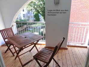 A wooden deck with a table and chairs, a white wall, and a view of a building through a glass railing. - Stralsund Altstadt, 2-Zi-Wohnung mit Loggia (Stralsund)