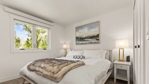A bedroom with a large bed, two bedside tables with lamps, a painting on the wall, and a window with a view of trees.