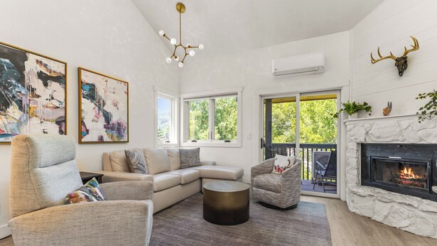 A modern living room with a sectional sofa, a round coffee table, and a fireplace. There are two large windows providing natural light and a view of greenery outside.