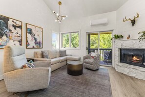 A modern living room with a sectional sofa, a round coffee table, and a fireplace. There are two large windows providing natural light and a view of greenery outside. - Minutes from Town & Slopes | Hot Tub |Private W/D | On Bus Route (Steamboat Springs)