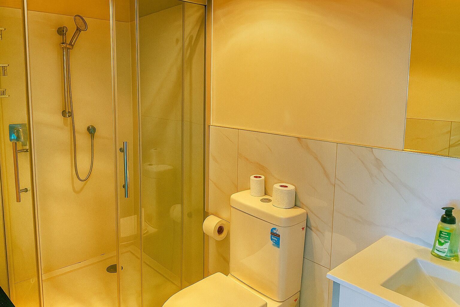 Un baño con ducha de vidrio, inodoro y lavamanos con dispensador de jabón.