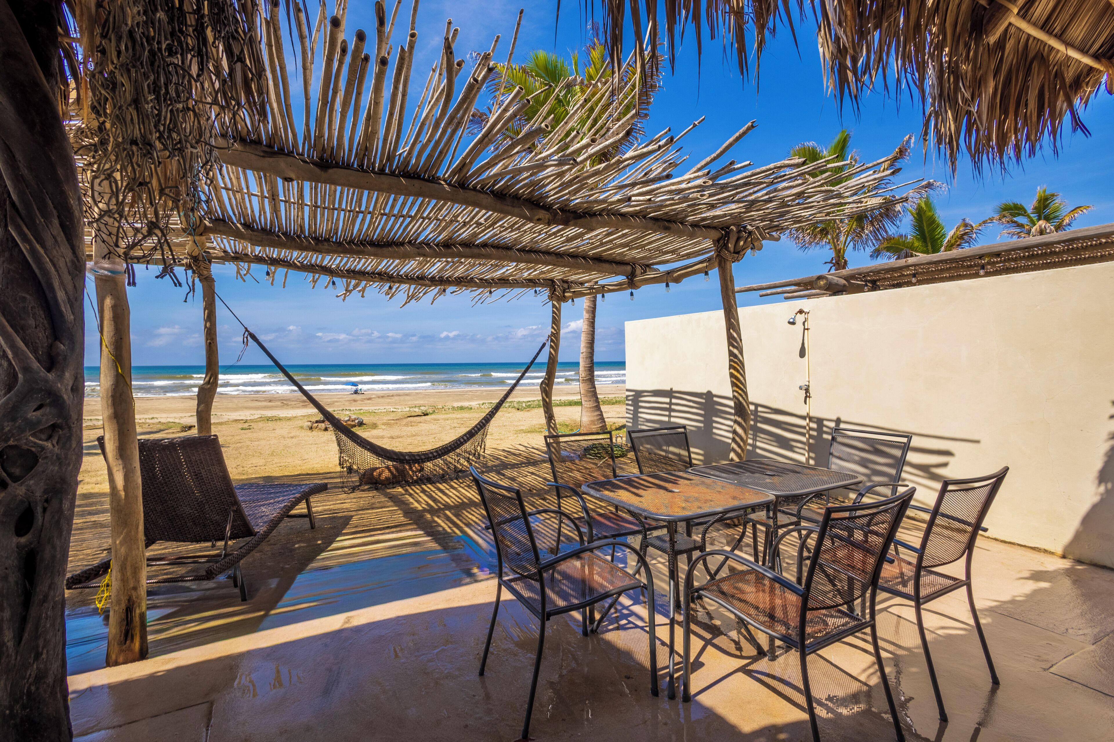 Un patio frente a la playa con techo de paja, una hamaca y una mesa con sillas.