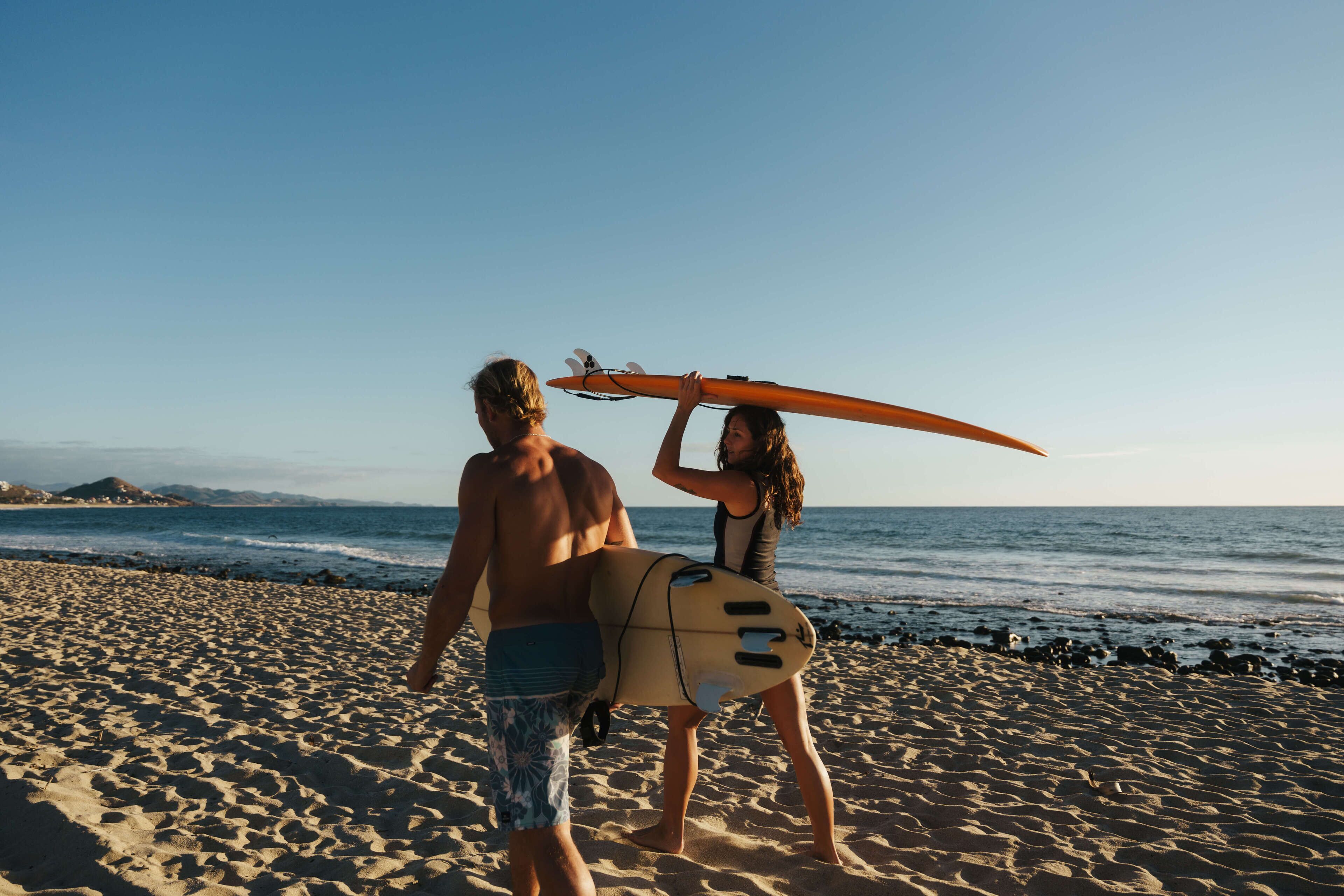 Escena de playa con dos personas caminando, una de ellas lleva una tabla de surf.