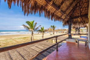 A beach view from a thatched-roof structure with a tiled floor, palm trees, and a distant building.