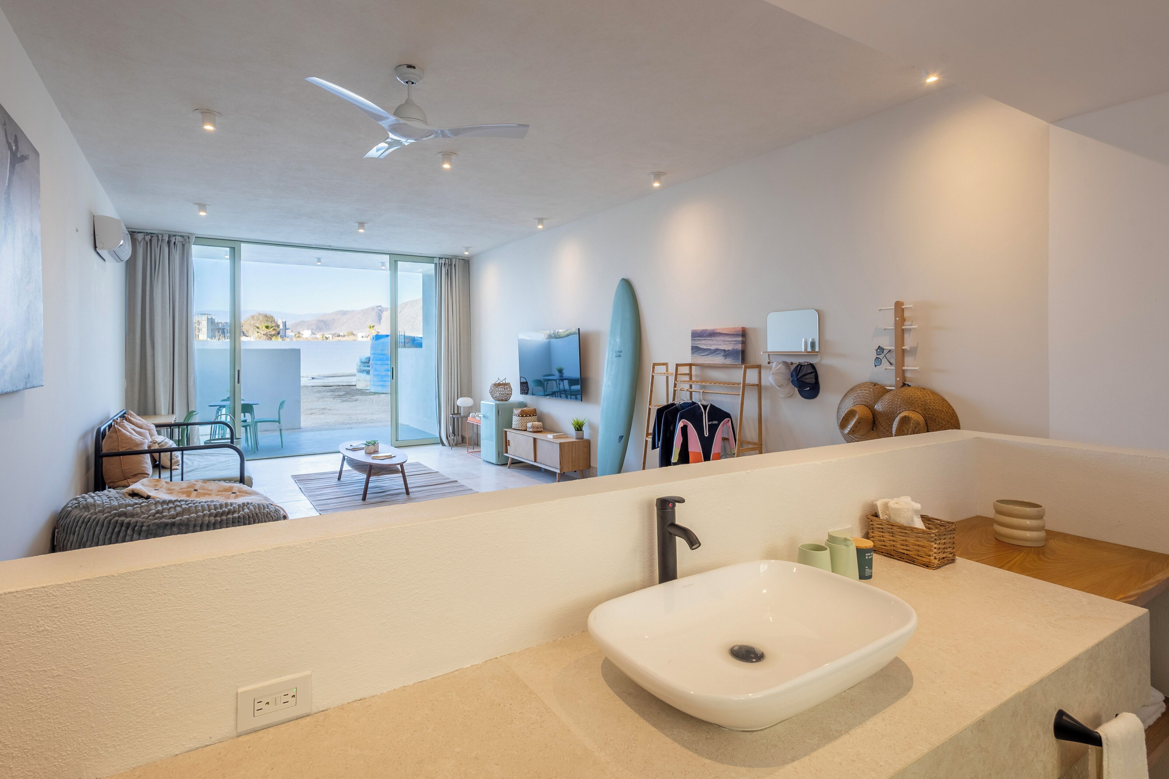 A modern bathroom with a large white sink, a ceiling fan, and a view of the living area with a sofa and a surfboard.