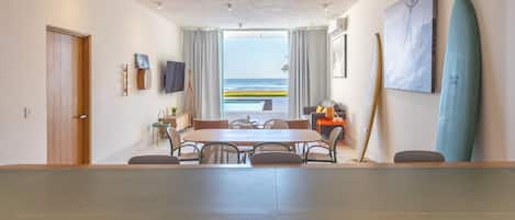 A spacious room with a large dining table, a view of the ocean, and surfboards on the wall.