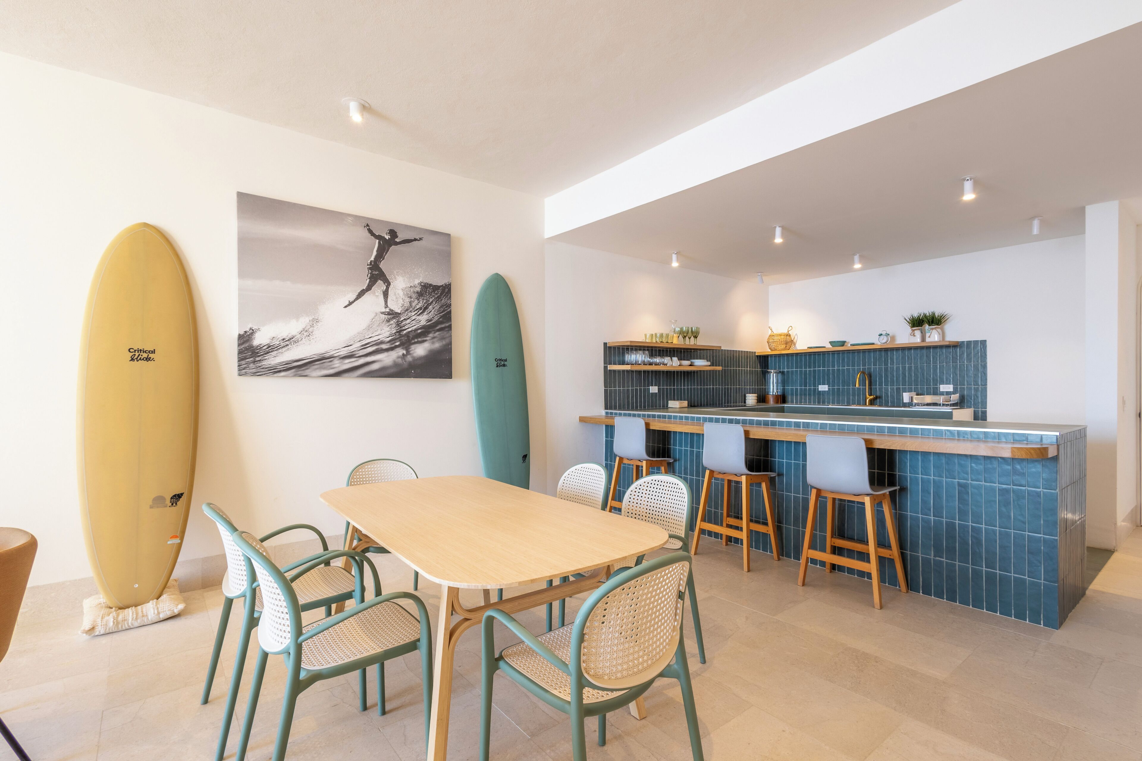 Une cuisine de taille réduite avec un plan de travail bleu recouvert de carreaux, une table à manger en bois accompagnée de quatre chaises, et des planches de surf accrochées au mur.