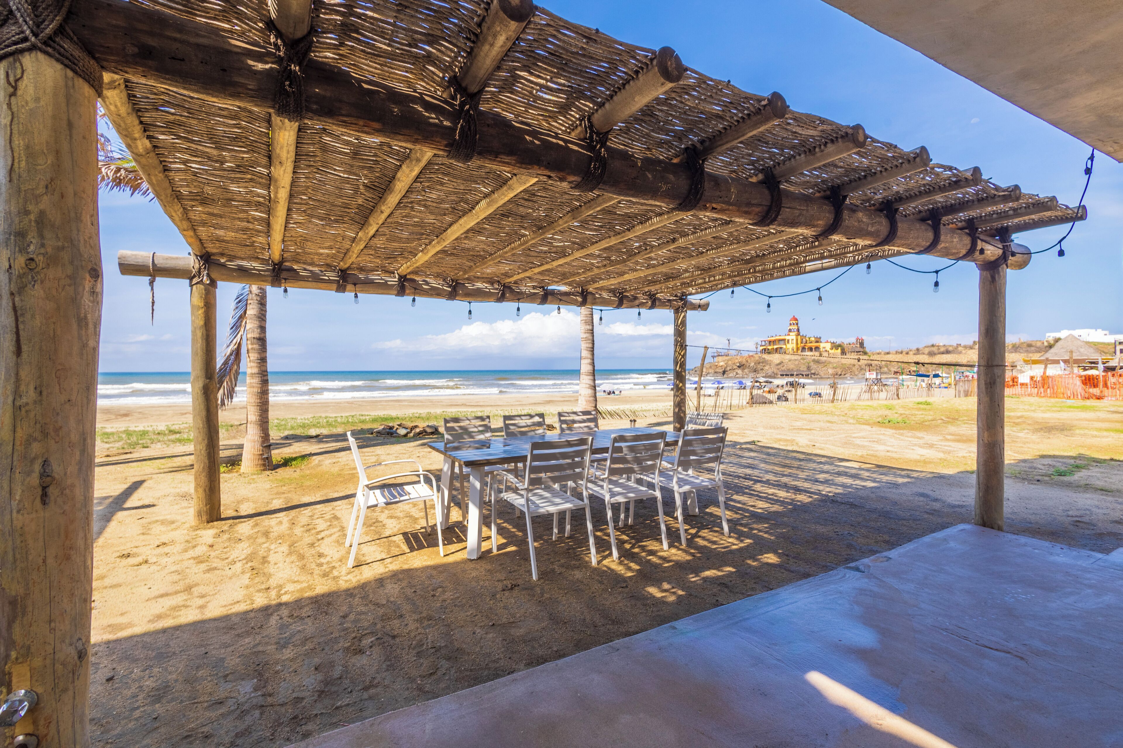 A beachside patio with a thatched roof, white metal chairs, and a view of the ocean.