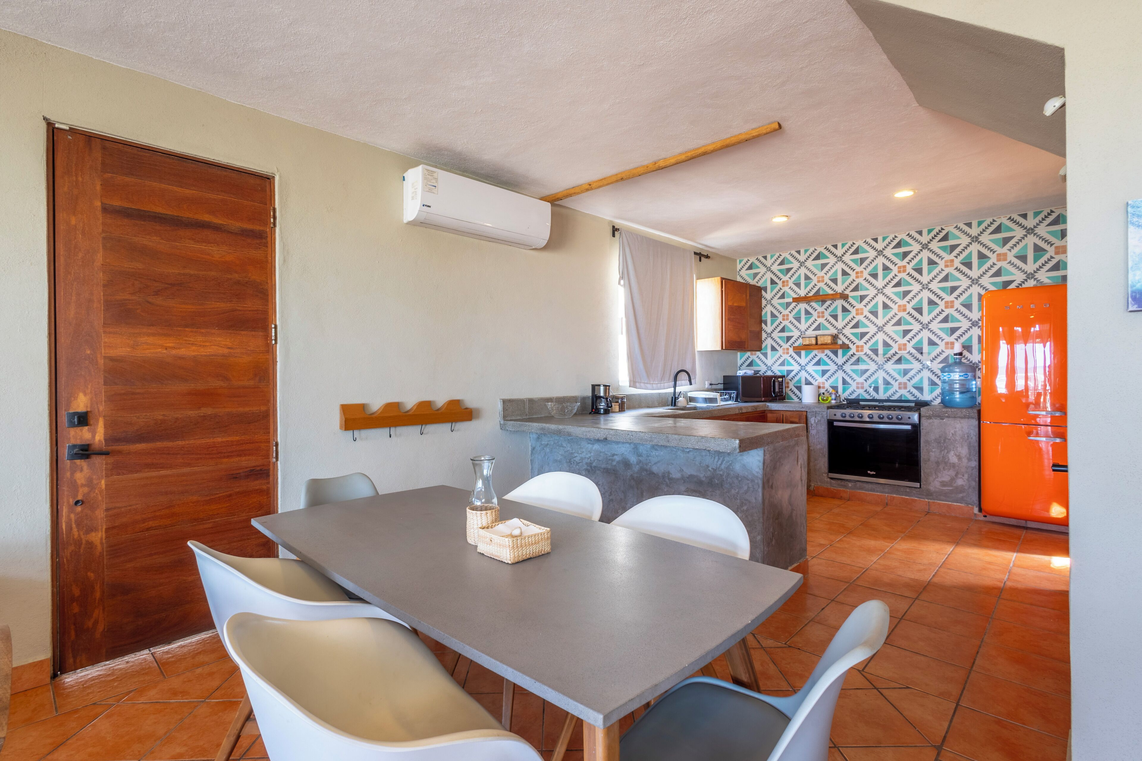 A compact kitchen with a dining area, featuring a wooden door, a small island with a sink, and a tiled backsplash.