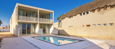 Una casa moderna de dos pisos con piscina, ventanas amplias y una estructura con techo de paja.
