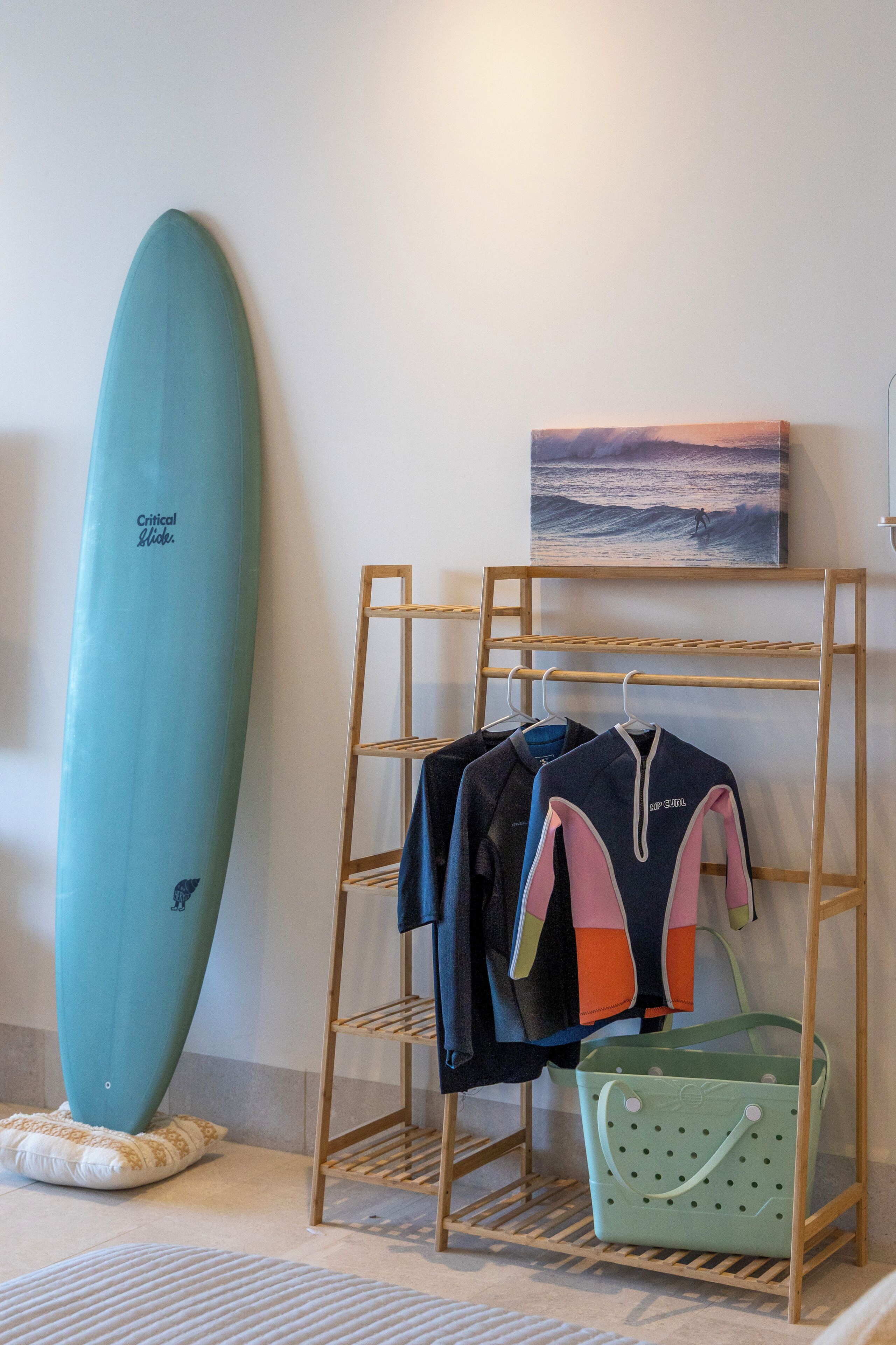 A surfboard, a rack with wetsuits, and a painting of a surfer on a wave.