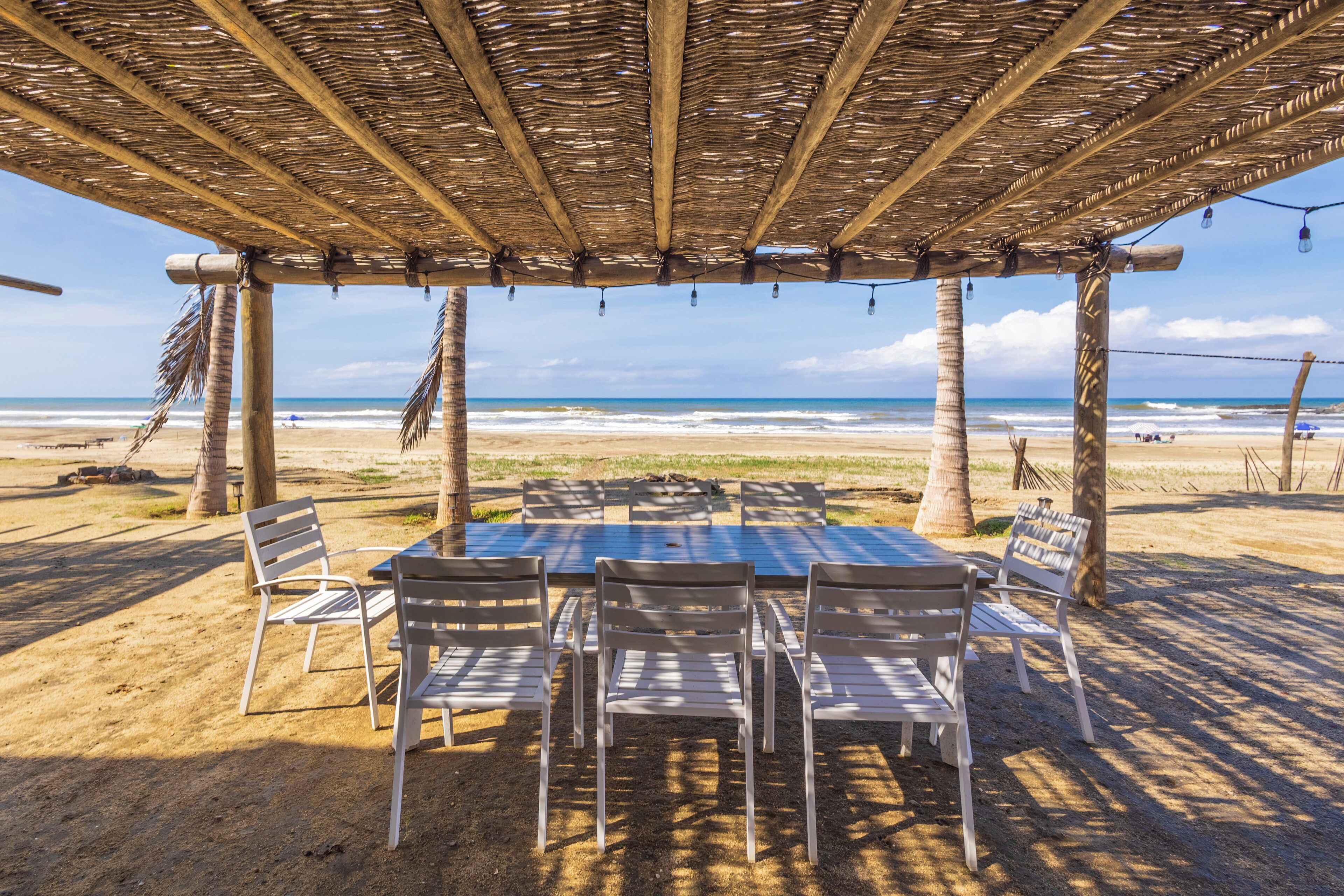 A beachside outdoor seating area with a table and chairs under a thatched roof, overlooking the ocean.