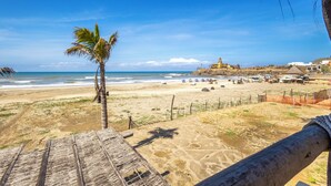 A beach view with a thatched roof structure, sandy beach, palm trees, and a distant building. - Costa Cerritos Beach Front Property (El Pescadero)