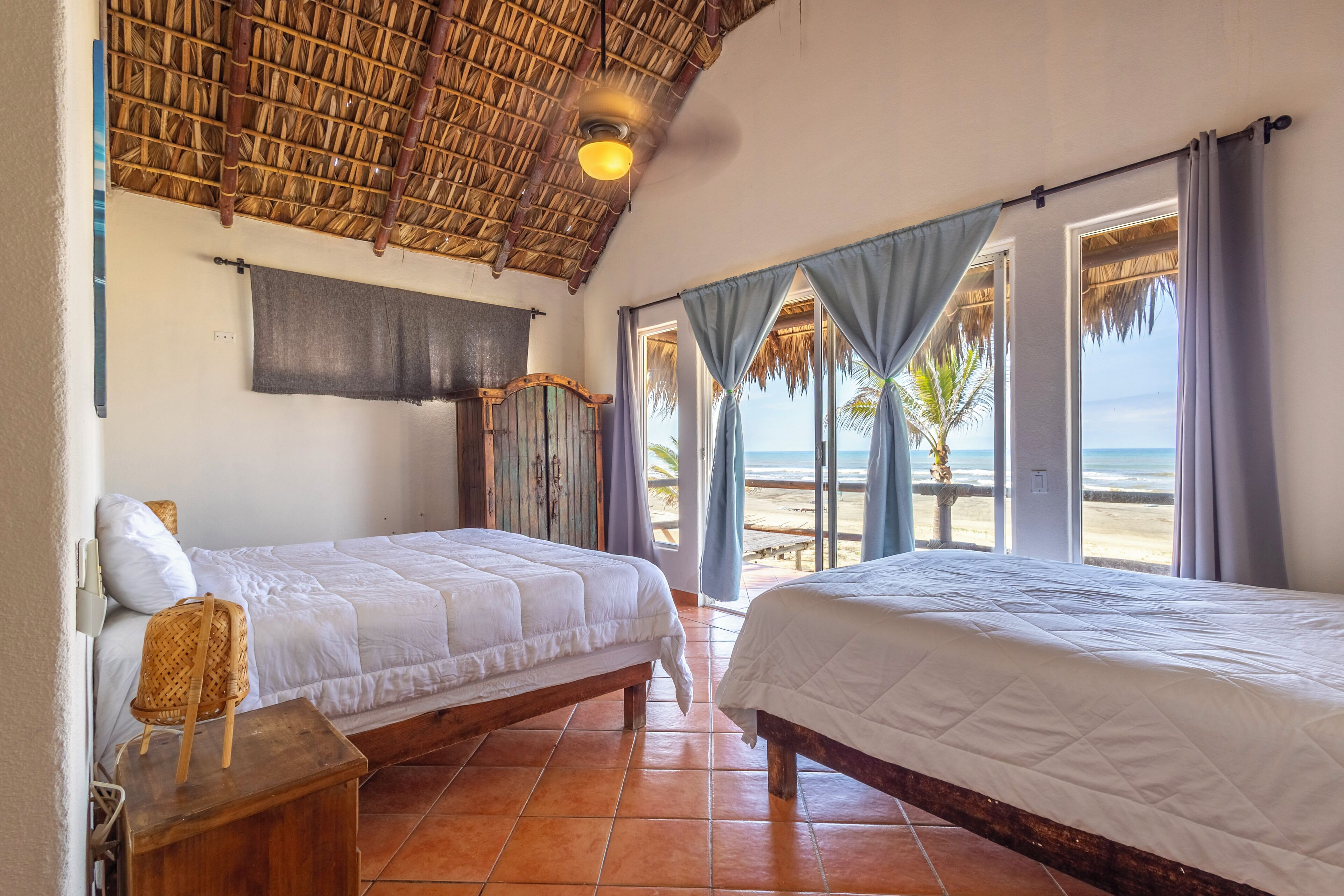 A room with two beds, a thatched ceiling, and a view of the beach.