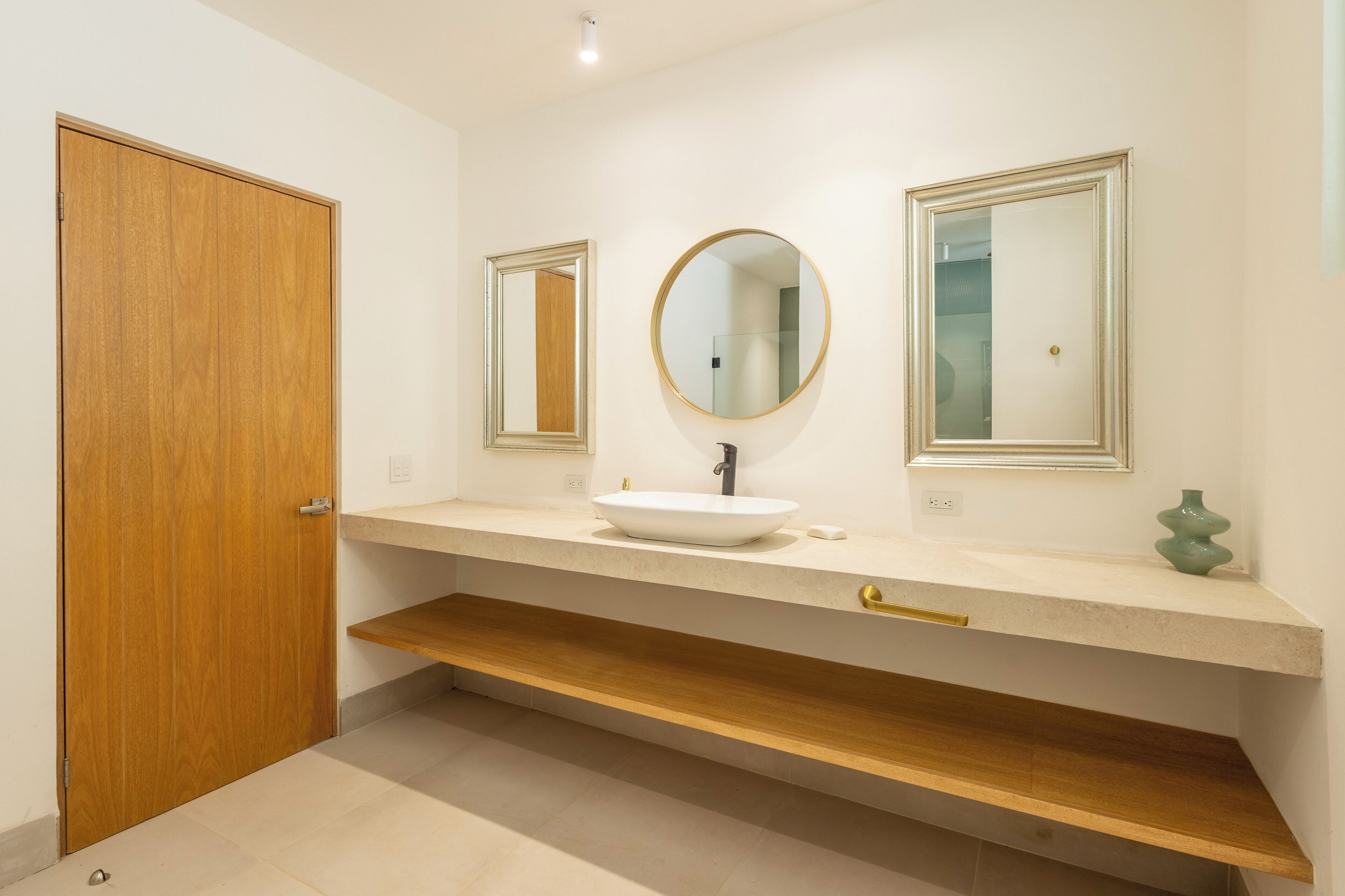 Un bany modern amb una porta de fusta, un lavabo blanc, un mirall rodó i un vas petit a la superfície.
