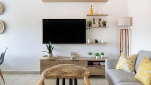 En stue med et fladskærms-tv på væggen, en grå sofa med gule puder, et træbord og et lille sofabord med en lampe.