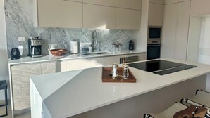 Une cuisine moderne avec un dosseret en marbre, des meubles blancs, un four encastré et un grand îlot central doté d’une machine à café et de salières.