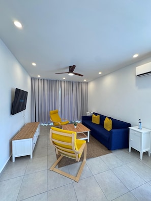 A modern living room with a wooden coffee table, yellow chairs, a blue sofa, a ceiling fan, recessed lighting, and a flat-screen TV mounted on the wall.